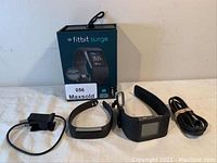 Lot of two Fitbit watches, one Surge with original box and USB charger, second smaller Fitbit fitness band with USB charger, all items shown together.