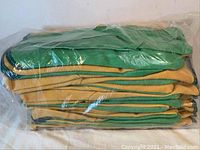 Stack of twelve pairs of green and yellow gardening gloves made of cloth and vinyl in plastic packaging.