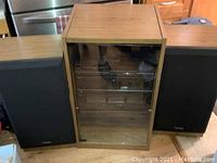 Pair of black front Technics mid-range tower acoustic speakers flanking a wooden stereo stand with glass display door.