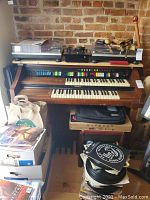 Front view of a wooden Lowrey electric organ with dual manuals, colorful switches, and pedalboard. Accessories including vinyl records, bags, and cases are shown around the organ.