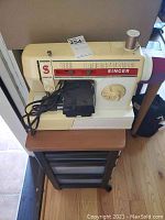 Front and left side view of cream-colored vintage Singer sewing machine with power cord and foot pedal, sitting on a small wooden table.