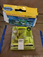Photo of the yellow plastic carry case with various drill bits, screwdriver bits, and scrubbing brush attachments, laid in front of the original box with visible damage.