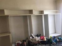 Photo showing three white shelving units against a wall above a cluttered table, units have open shelving compartments