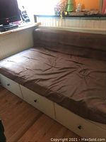 Daybed with brown cushion seen from angled front-left side, showing cushion and storage drawers under seat.