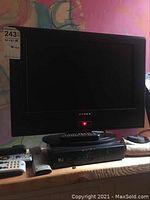 Black 26 inch Dynex television on a wooden surface with a red power indicator light on, accompanied by remotes and electronics underneath.