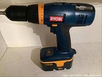 Blue and black Ryobi cordless drill with orange accents and an 18V battery attached placed on a white shelf.