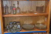 Shelf display showing various glass items: two glass angels, a glass bell, Coca Cola bottle, glass bowls and plates, glass jar mugs with lids, and a metal tray beneath.