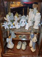 Full display of 14 ceramic figurines including angels playing instruments, cherub, standing human figures, and ceramic baby shoes on glass shelf.