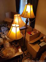 Photo of three matching floor lamps including the tallest at 62 inches, with fabric shades and decorative bases. Lamps are illuminated and pictured next to cardboard boxes.