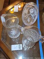 Overview of two clear crystal pitchers and one crystal etched basket on glass table surface
