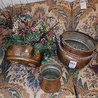 Photo shows three brass planters placed on a patterned fabric surface. One planter holds artificial plants and flowers, another has a hammered texture, and the smallest is plain with visible black stamped numbers.