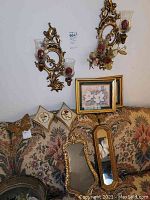 Photo showing two ornate gold-tone candle wall sconces with attached glass shades, two floral print picture frames, one ornate gold-tone mirror, and two butterfly wall decorations all displayed on a floral patterned fabric surface and wall.