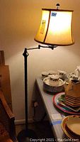 Full view of the metal swing arm floor lamp with beige fabric shade turned on, standing beside a table with dishes.