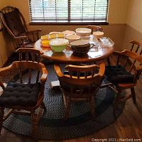 Solid oak round dining table with four matching solid oak chairs around it. Chairs have black cushioned seats. Table top has various kitchen bowls and containers on it.