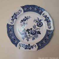 Blue and white ceramic wall plate with floral design and diamond-patterned border shown hanging on the wall with plate hangers