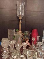 Full view of assorted glass candle holders including tall hurricane style and several smaller votive and stick holders, along with red mini lamp shades.