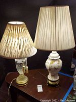 Two table lamps on dark wood table; left lamp clear cut glass base with diamond pattern and tassel pull chain, right lamp ceramic base with painted butterflies and plain pleated shade.
