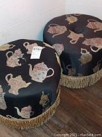 Pair of stools, round cushioned seats covered in black fabric with printed teapots and gold fringe trim along the base.