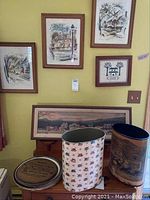 Four framed watercolour prints by John Hayman on the wall, plus a panoramic cityscape print framed below, with two decorative tin trash cans and four metal platters on a table
