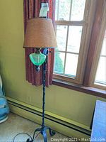 Full view of vintage floor lamp showing twisted wrought iron stand, green glass accent, and beige fabric lampshade near window.