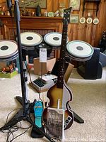 Full view of drum set with pads and stands, guitar controller, microphone, and game case on carpeted floor.