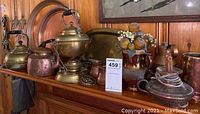 Photo showing brass and copper teapots, platters, creamers arranged on wood surface with a wood wall in the background.