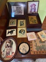 Overview of all framed art prints on a wooden table, featuring dog-themed prints, a Currier & Ives print, and a vintage thermometer print.