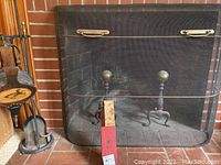 Photo showing black metal fireplace screen with mesh, brass-tone handles, and two andirons with brass tops behind it.
