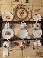 Photo of two wooden shelves mounted on a wall with collectible ceramic cups, saucers, decorative plates, and glass salt and pepper shakers arranged on them. Cups have various designs including animals and floral patterns. Condition shows signs of crazing and wear.