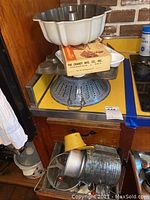 Photo of vintage kitchen items piled including bundt pan, metal colanders, flour sifter, baking pans with some original packaging.