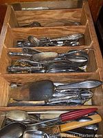 Overview of assorted flatware and serving pieces neatly arranged in a wooden divided organizer tray, showing various knives, spoons, and forks.