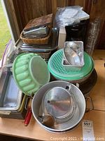 Photo of assorted bakeware including roasting pans, baking trays, green bundt mold, plastic cake pans with lids, and a wicker basket filled with various items.