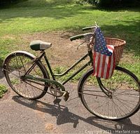 Side view of green Schwinn ladies bicycle with wicker basket and American flag on handlebar, showing weathering, oxidation, and flat tires.