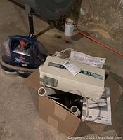 Oreck XL portable vacuum in beige with attachments in a box along with manuals and accessories, situated on a basement floor.
