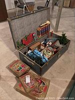 Vintage fabric-lined trunk open showing assorted Christmas decorations including boxes of bulbs, miniature truck, candles, small figurines, and greenery.