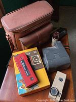 Photo of multiple vintage cameras and accessories including Kodak cameras, Argus 35mm with leather bag, grey Polaroid camera, and original Kodak Disc 8000 box.