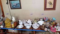 Wide photo showing an assortment of Belleek porcelain items, transferware, hobnail milk glass, dog figurines, mantle clocks, crystal and kitchen decor items arranged on a table against a wall with some framed pictures.