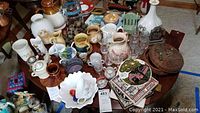 Overview of table with varied ceramics, glassware, pottery, and collectibles including pitchers, bowls, figurines, and glassware