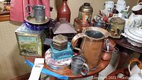 Photo showing an assembled assortment of vintage kitchen items including a large decorative tin box, a copper pitcher, brass mugs, and a blue tobacco tin.