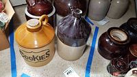 Photo shows collection of vintage crockery including a tan ceramic cookie jar labeled 'COOKIES' and several brown glazed stoneware jugs with various shapes and sizes, placed on floor with other similar jars.