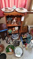 Photo of wooden display shelves featuring dog figurines, collector plates on shelves and floor, tin bank, and assorted collectibles.