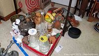 Wide shot showing collection of glass jars, holiday novelties, faux fruit in wooden bowls, plush toys, cast iron skillet, metal lantern, ceramics, and other kitchen goods grouped on floor.