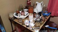 Photo showing a wide assortment of ceramic teapots, pitchers, brass figurines, glassware, and various kitchen and home decor items arranged on a table.