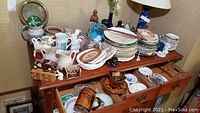 Wide view of wooden side table covered with stacks of decorative china plates, ceramic pitchers, animal figurines, miniature model cars, brown pottery mugs, glassware, and various vintage collectible items.