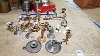 Full view of assorted brass, silver-tone, ceramic, filigree, and novelty candlesticks arranged on carpeted floor