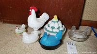 Five hen and rooster decorative dishes and figurines arranged together on floor showing different colors and styles.