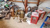 Wide photo showing the majority of metal items on the floor, including lanterns, coffee pots, dog doorstop, sad irons, and other metalwares.