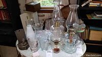 Multiple clear glass oil lamps with brass fittings, hurricane lamp chimneys, various glassware, and barware on round table.