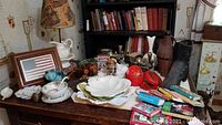 Photo of assorted lot items on a wooden table including marble locomotive bookends, cast iron boot figurine, red lacquer coasters, mini books, porcelain plates, dog figurine and a lamp with a patterned shade.