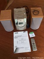 Photo showing the JVC FS-V5 receiver with CD player, two wooden speakers, user manual, and remote control arranged on a wooden surface.
